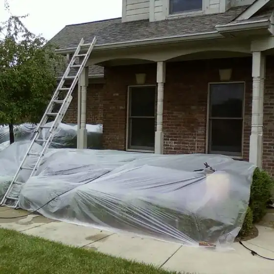 A brick house with plastic sheeting draped over the shrubs and a ladder leaned against the porch roofline before painting