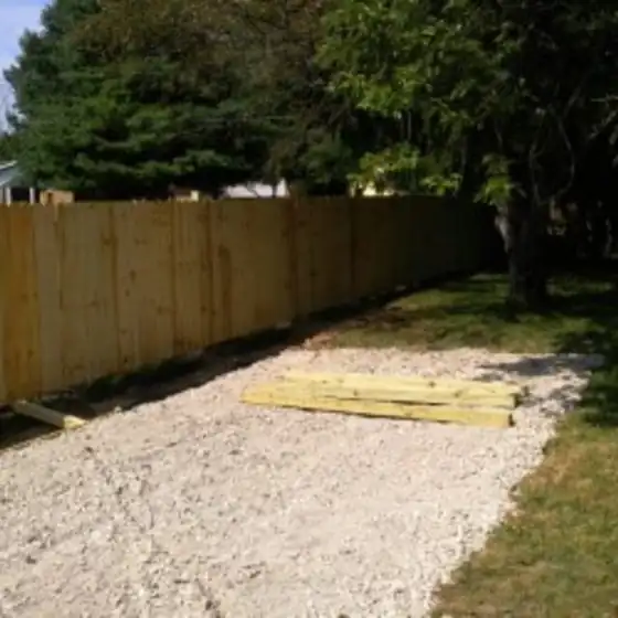 New wood privacy fence along a lawn, with a gravel strip and a stack of lumber in front of it
