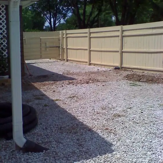Tan-painted wood privacy fence with a gate, a gravel yard in front and a downspout at the left