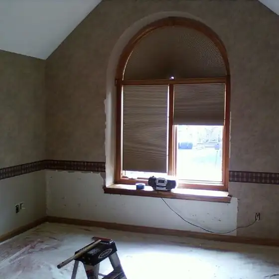 A bedroom with an arched window, wallpaper stripped back to bare wall below the border and drop cloths on the floor