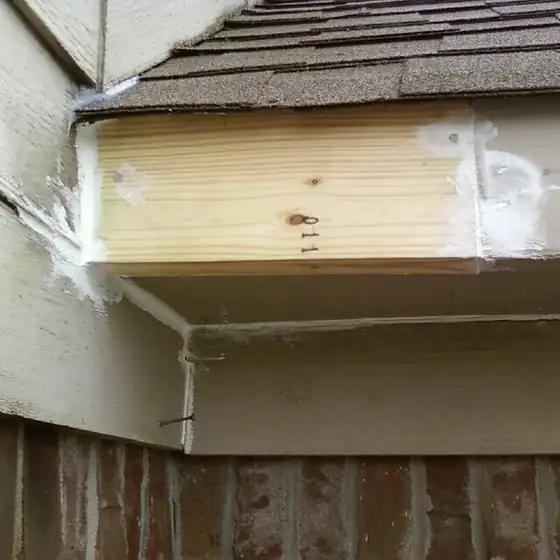 A fresh pine board fitted at the corner of a roofline above lap siding and brick, with patched edges either side