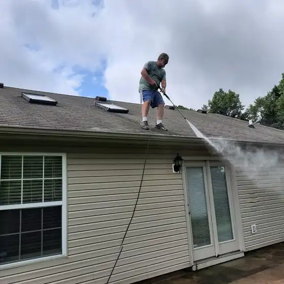 Worker pressure washing a shingle roof with a wand above a tan-sided house